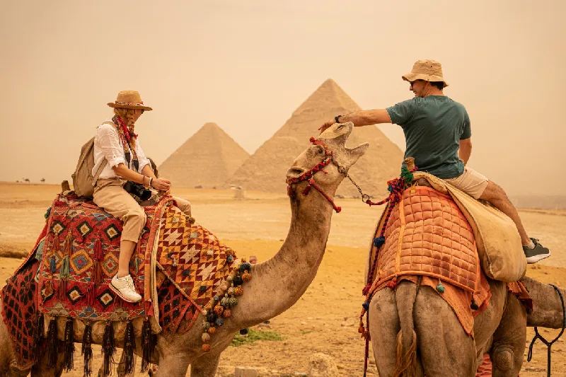 Turistas pasean en camello cerca de las pirámides de Giza, disfrutando una experiencia cultural única bajo un cielo brumoso.