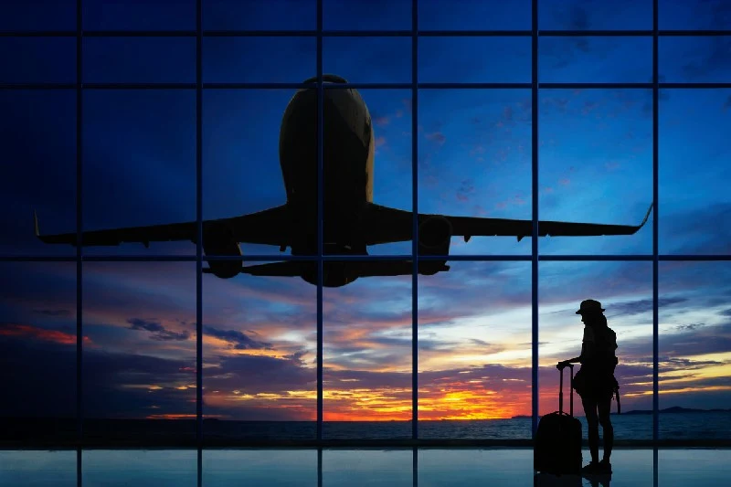 Persona con maleta observando un avión despegar al atardecer desde la ventana del aeropuerto