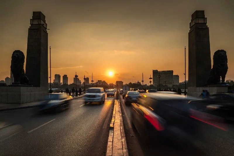 Vista de coches circulando por un puente en El Cairo durante el atardecer, con edificios al fondo y el cielo naranja.