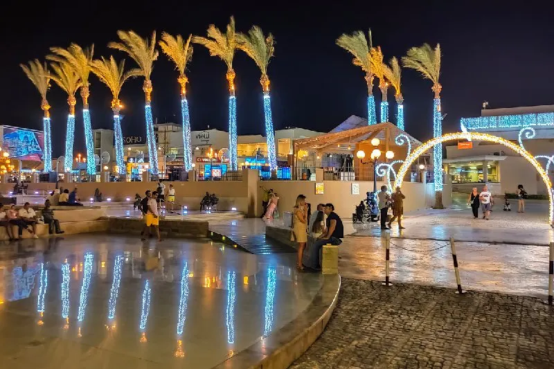 Personas paseando por SOHO Square en Sharm El Sheij con palmeras iluminadas de noche.