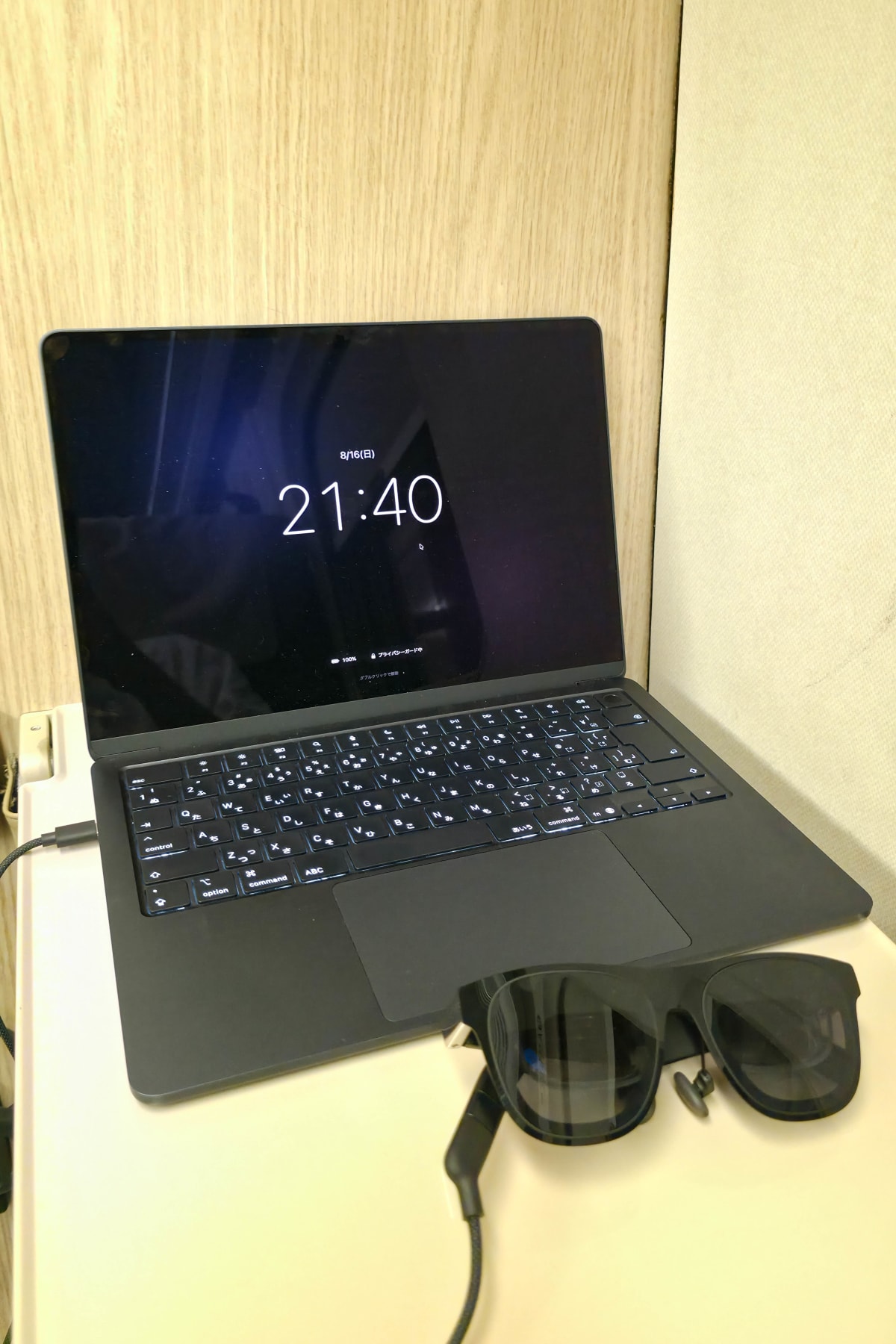 新幹線のテーブルに置かれたMacBook。本体の画面は時計だけのカバー画面で覆われ、日付と時刻、バッテリー残量、「ダブルクリックで解除」の文字だけが表示されている。手前にUSB-Cケーブルで繋がれたARグラスが置かれている