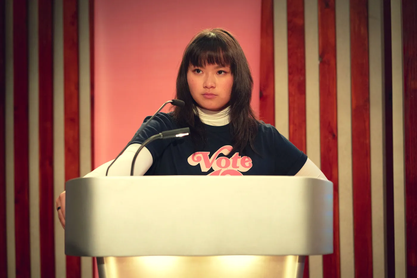 The Sex Education character O standing behind a lectern