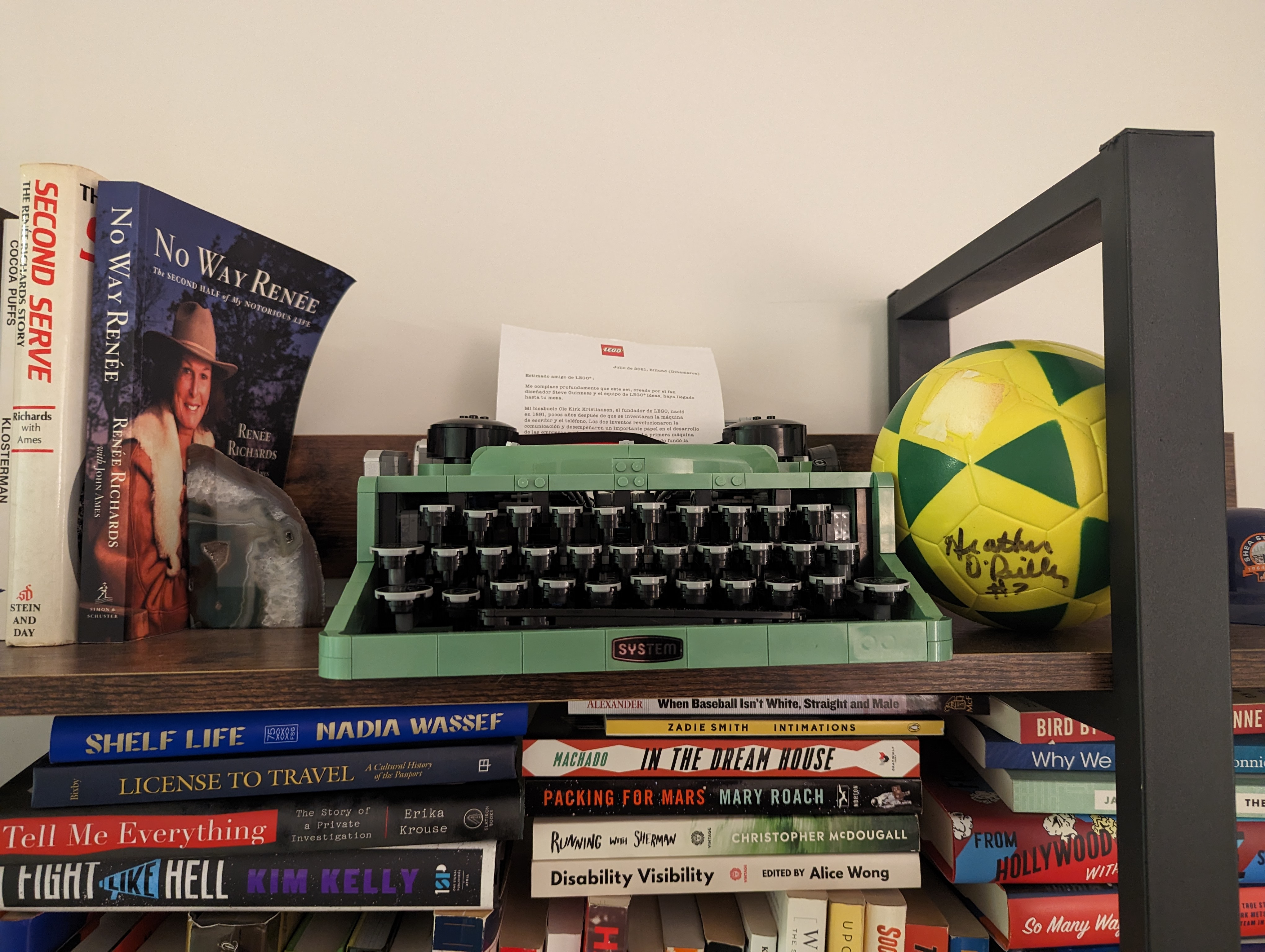 A green typewriter sitting on a bookshelf, surrounded by books and a small yellow autographed soccer ball
