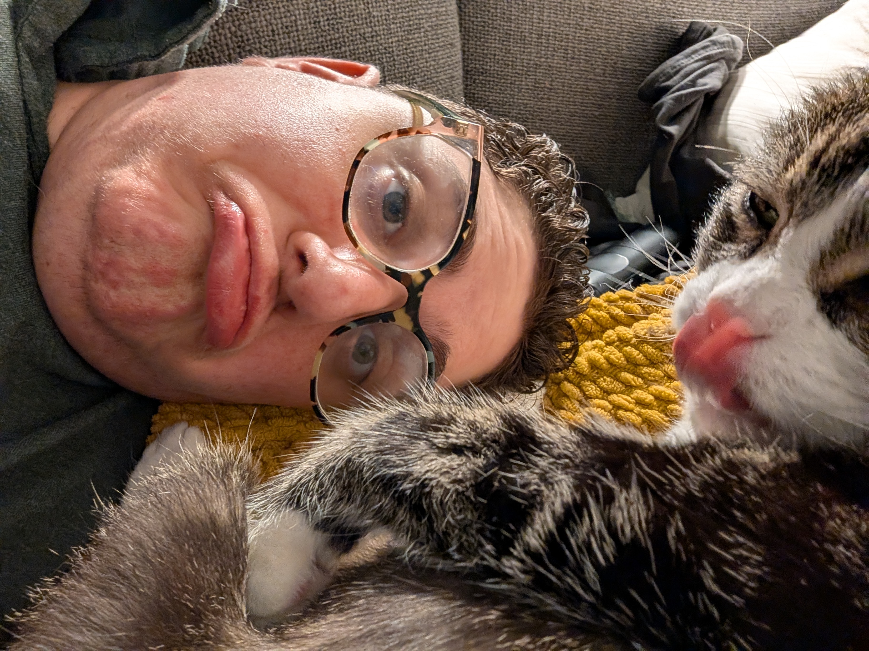 A selfie of me with my head sideways and my gray and white tabby cat with her tongue out
