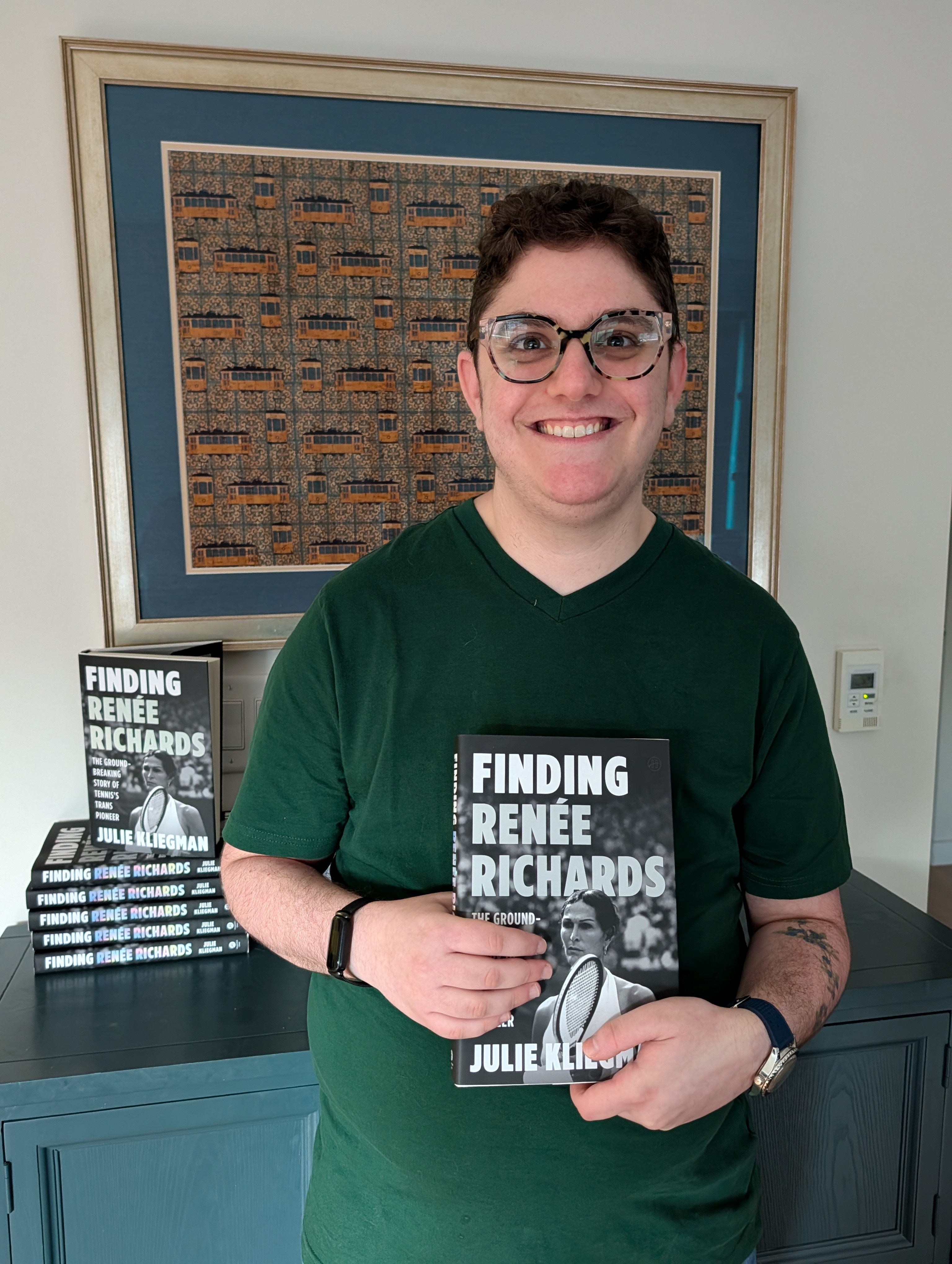I am wearing a forest-green shirt and holding a copy of the book Finding Renée Richards. I am standing in front of a blue console table and a piece of artwork on the wall. Behind me, there is a stack of other copies of Finding Renée Richards.