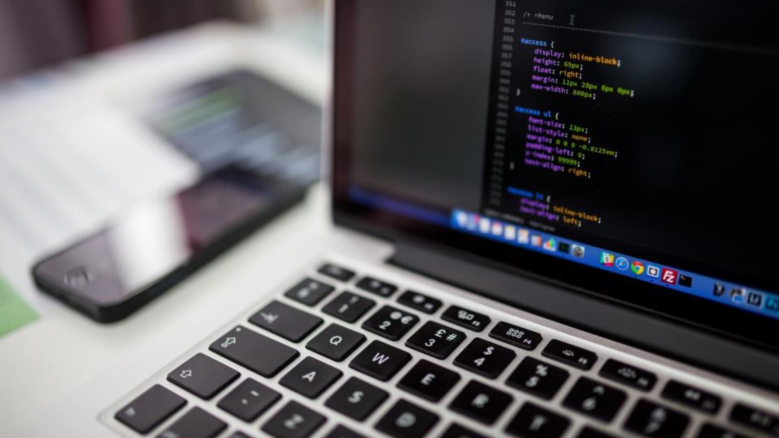 Portátil abierto mostrando código en un editor de texto sobre fondo oscuro, con un teléfono móvil al lado