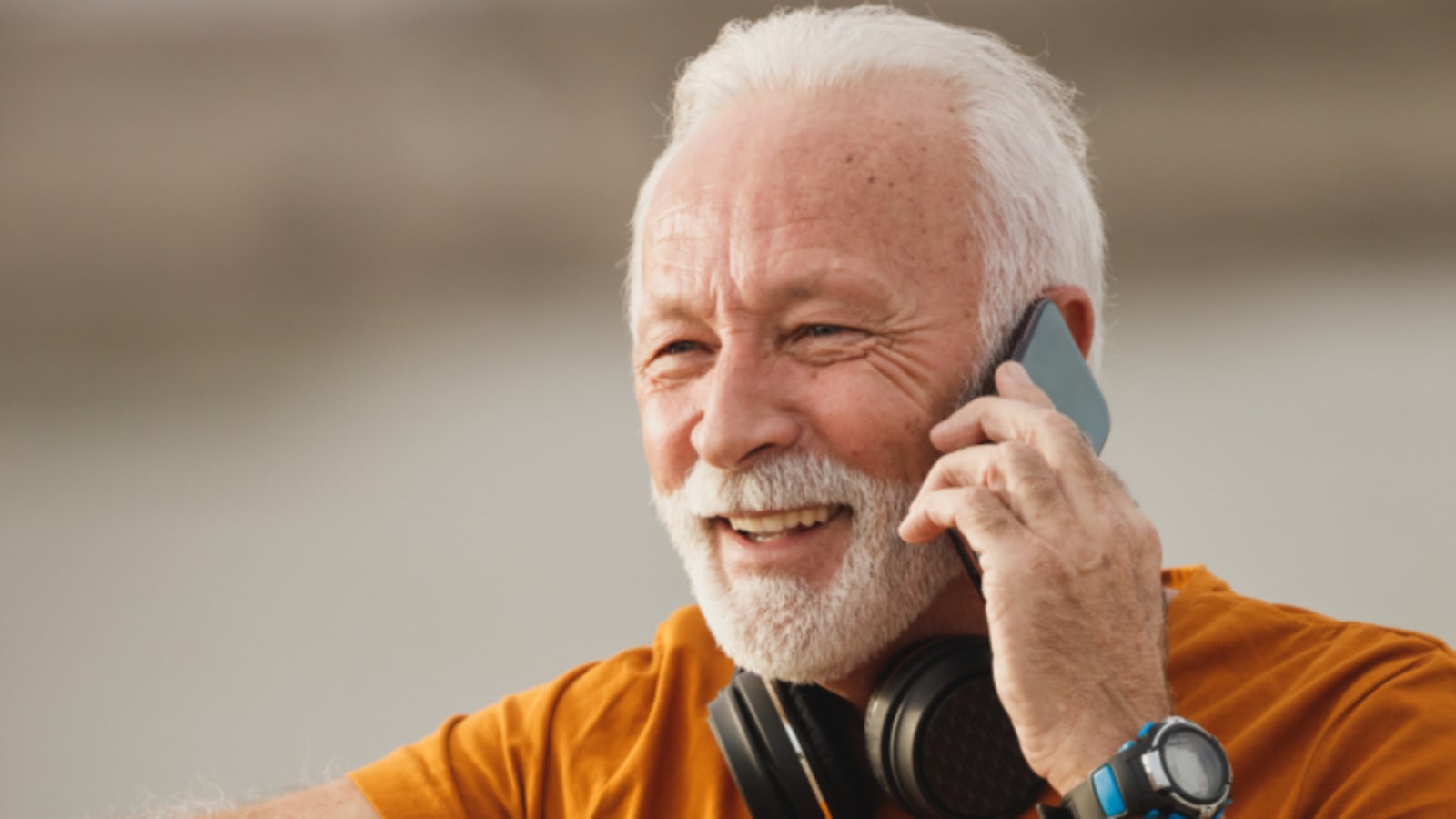 Hombre mayor sonriente hablando por un teléfono móvil con unos auriculares al cuello