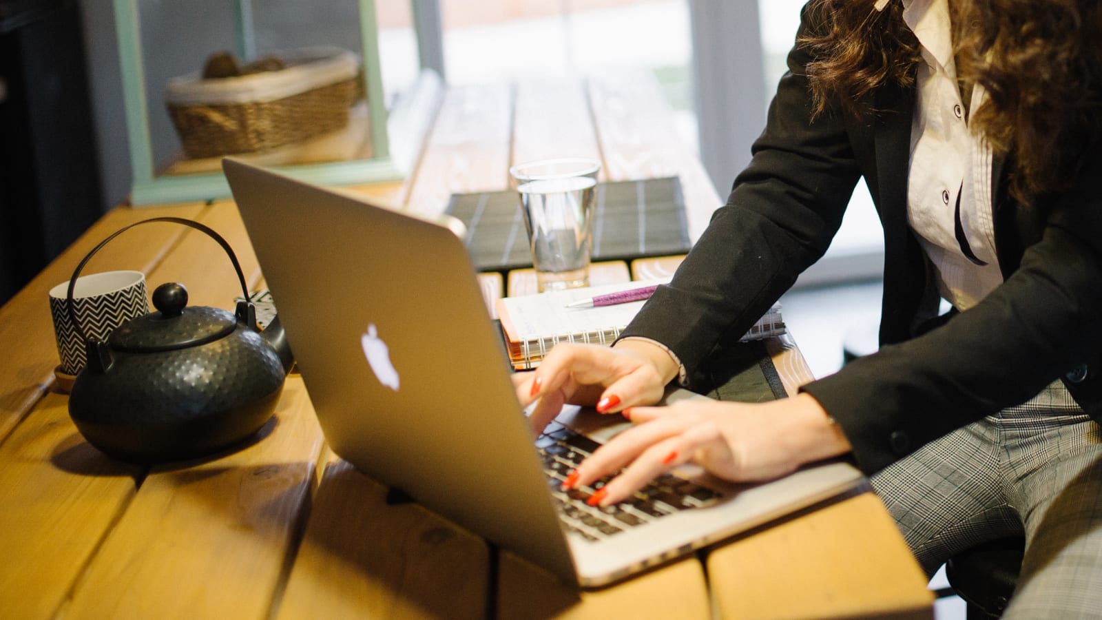 Una profesional escribe en el teclado de su portátil sobre una mesa de madera, con una libreta y una tetera al lado