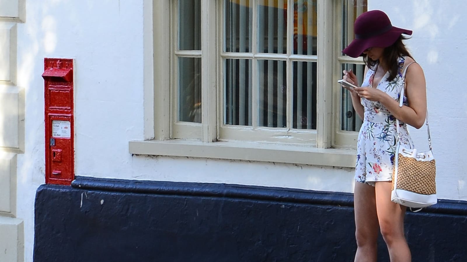 Buzón de correo rojo en la fachada de un edificio y, a unos metros, una mujer consultando su teléfono móvil en la calle