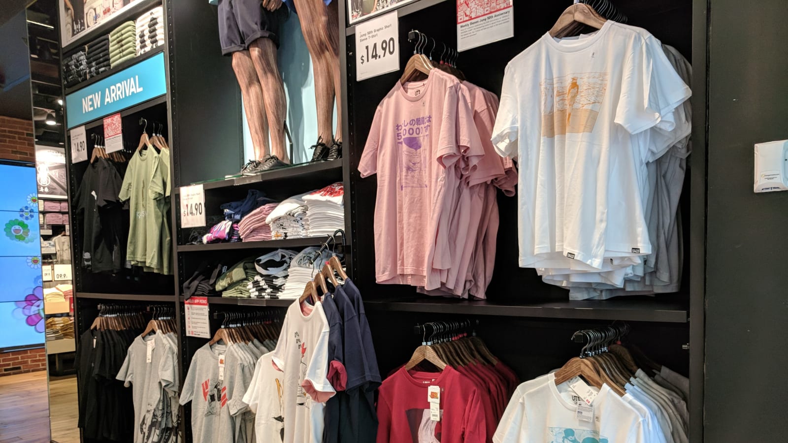 Interior de una tienda de ropa con estanterías y perchas de camisetas, con carteles de precio y un rótulo de novedades