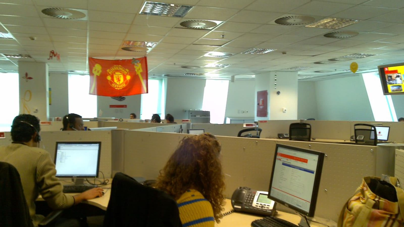 Sala de un centro de atención al cliente con puestos separados por mamparas, empleados con auriculares frente a sus pantallas