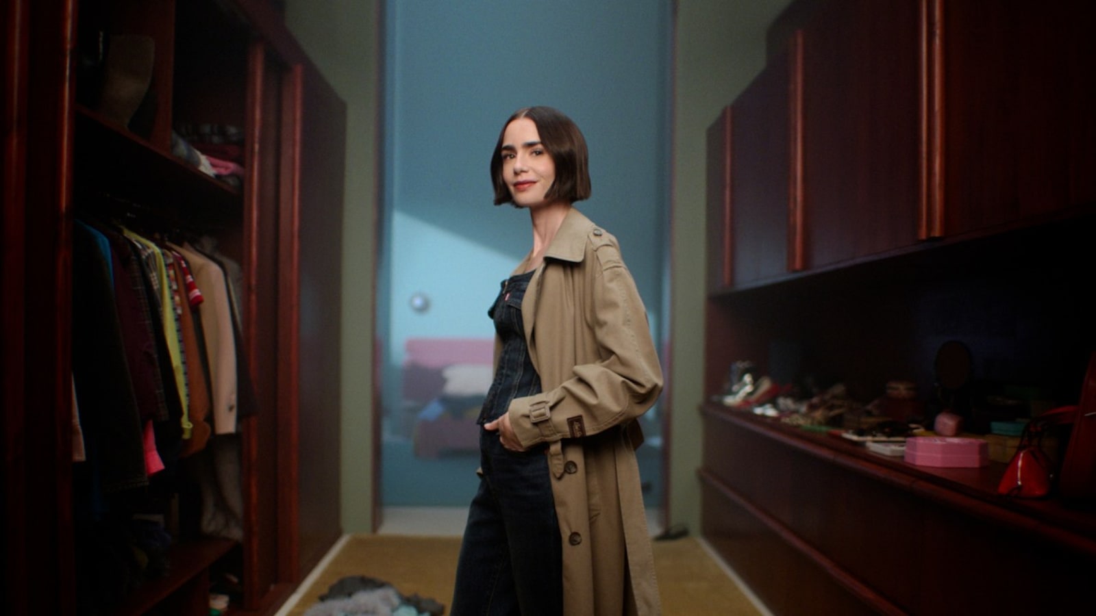 Lily Collins, con gabardina beige sobre un peto vaquero, de pie en un vestidor con armarios de madera llenos de ropa y zapatos