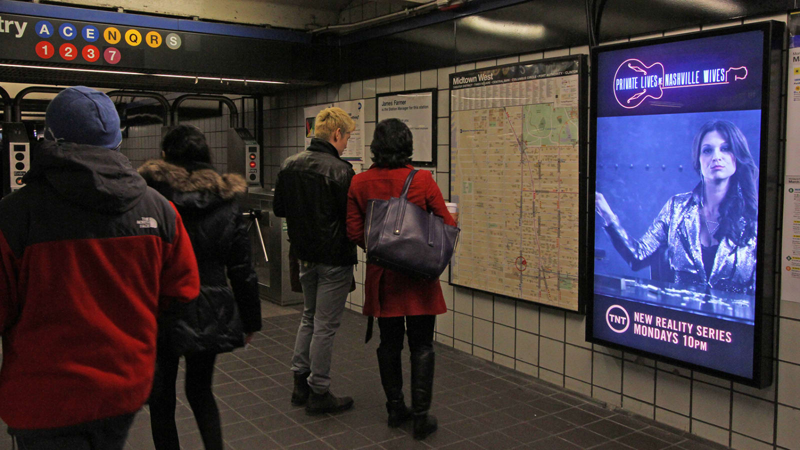 Pantalla publicitaria digital iluminada en el vestíbulo de una estación de metro, con varias personas caminando delante