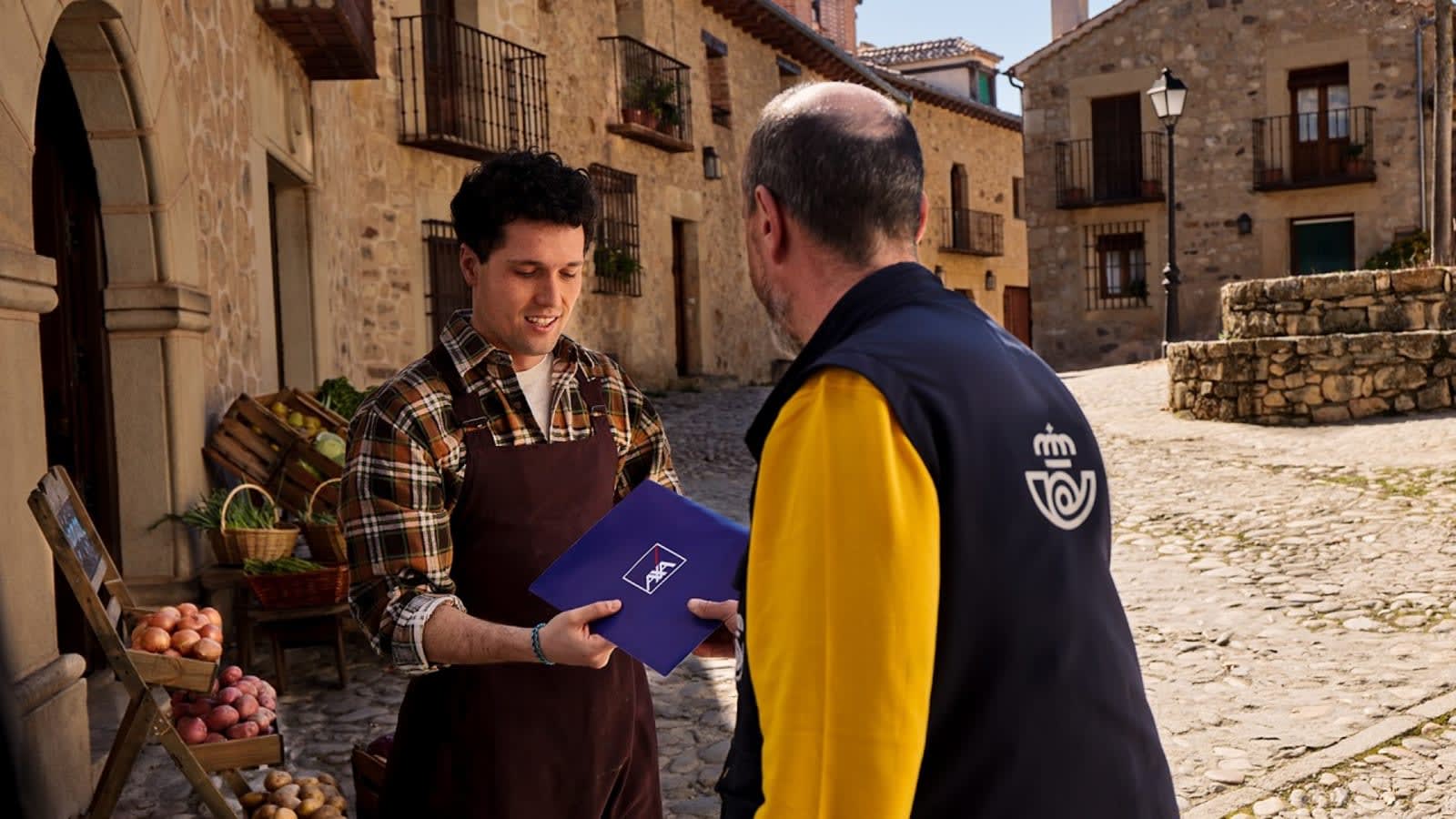 Un cartero de Correos entrega un sobre azul de AXA a un tendero en la calle empedrada de un pueblo de piedra