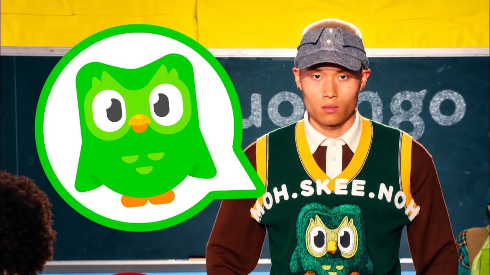 Modelo con gorra y chaleco de punto verde con el búho de Duolingo y la palabra MOH.SKEE.NOH, ante una pizarra de aula