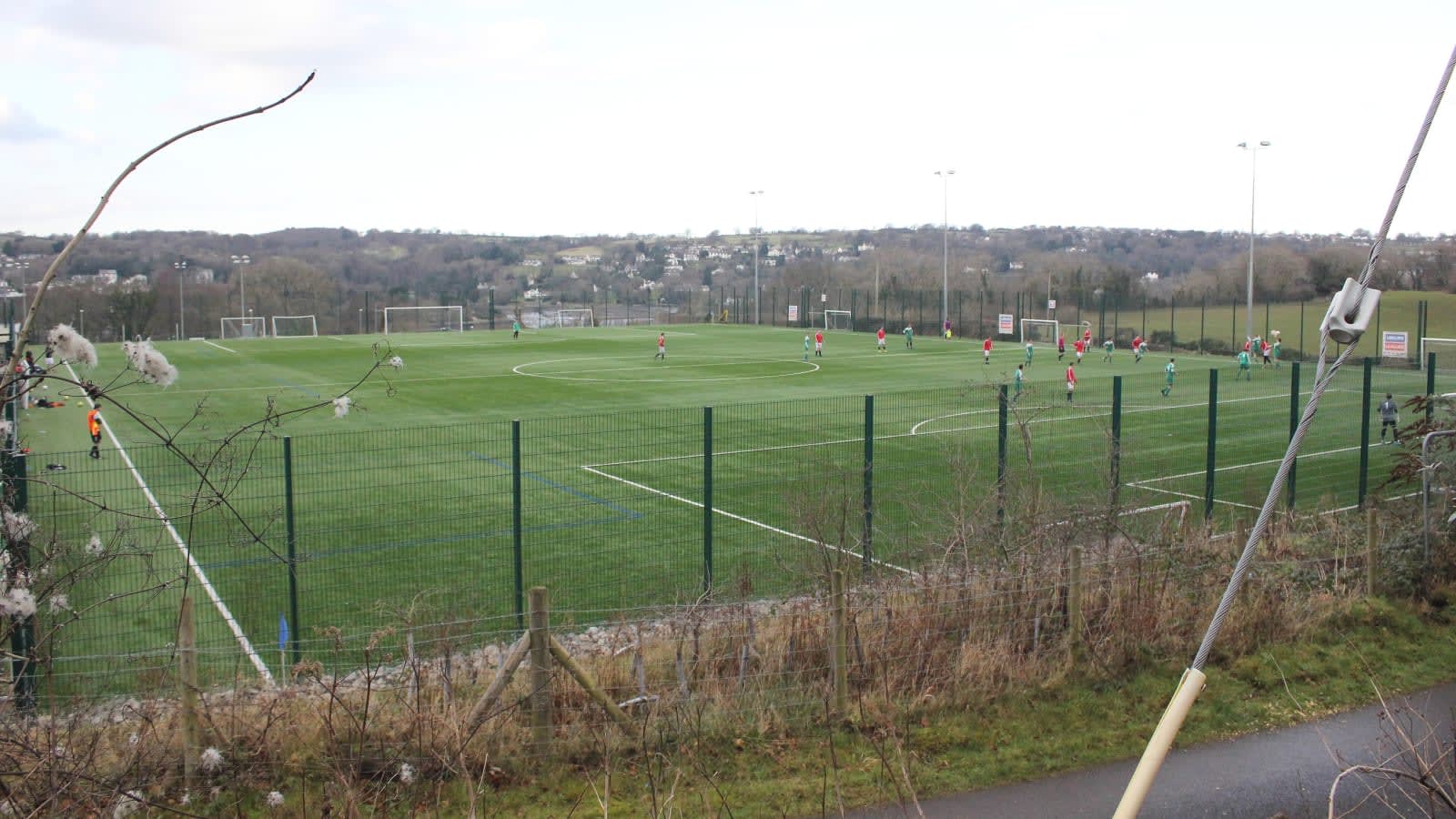 Campo de fútbol de hierba con un partido en juego, visto desde fuera de la valla, con casas y colinas al fondo