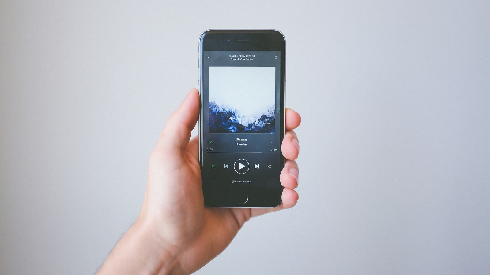 Una mano sostiene un móvil con la aplicación de Spotify reproduciendo una canción, sobre fondo gris