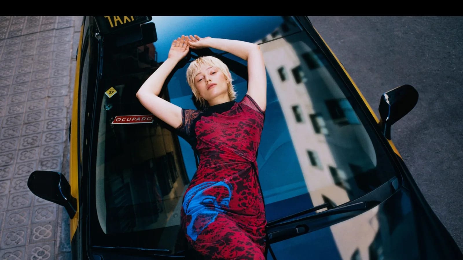 Una modelo con vestido rojo estampado posa tumbada sobre el capó de un taxi negro y amarillo de Barcelona con el cartel de ocupado, junto a una acera de panot