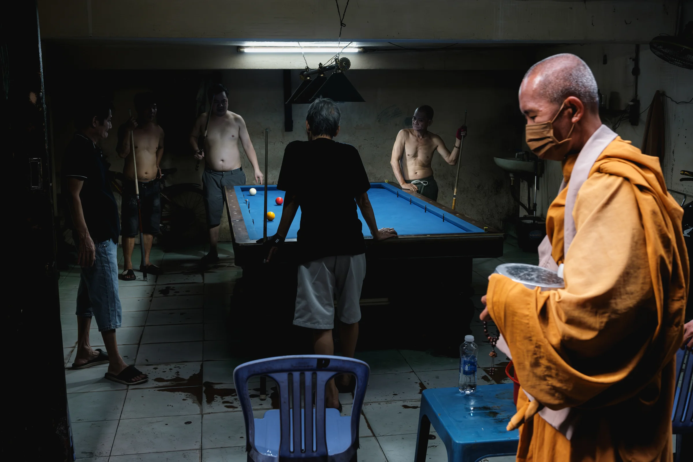 A saffron-robed, masked monk holding an alms bowl and prayer beads passes through a dim backstreet pool hall where shirtless men lean on cues around a blue-felt table — Adrien Jean.