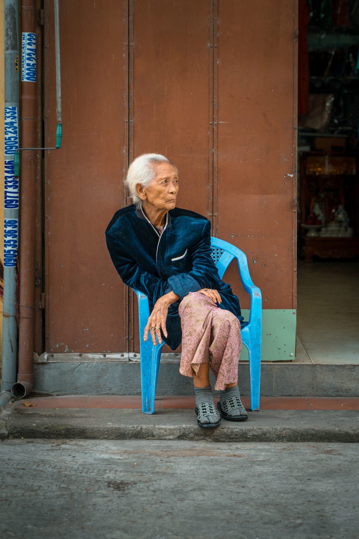An elderly white-haired woman in a dark velvet jacket and patterned trousers sits on a blue plastic chair on a doorstep, gazing to the side in front of a brown metal door — Pete Walls