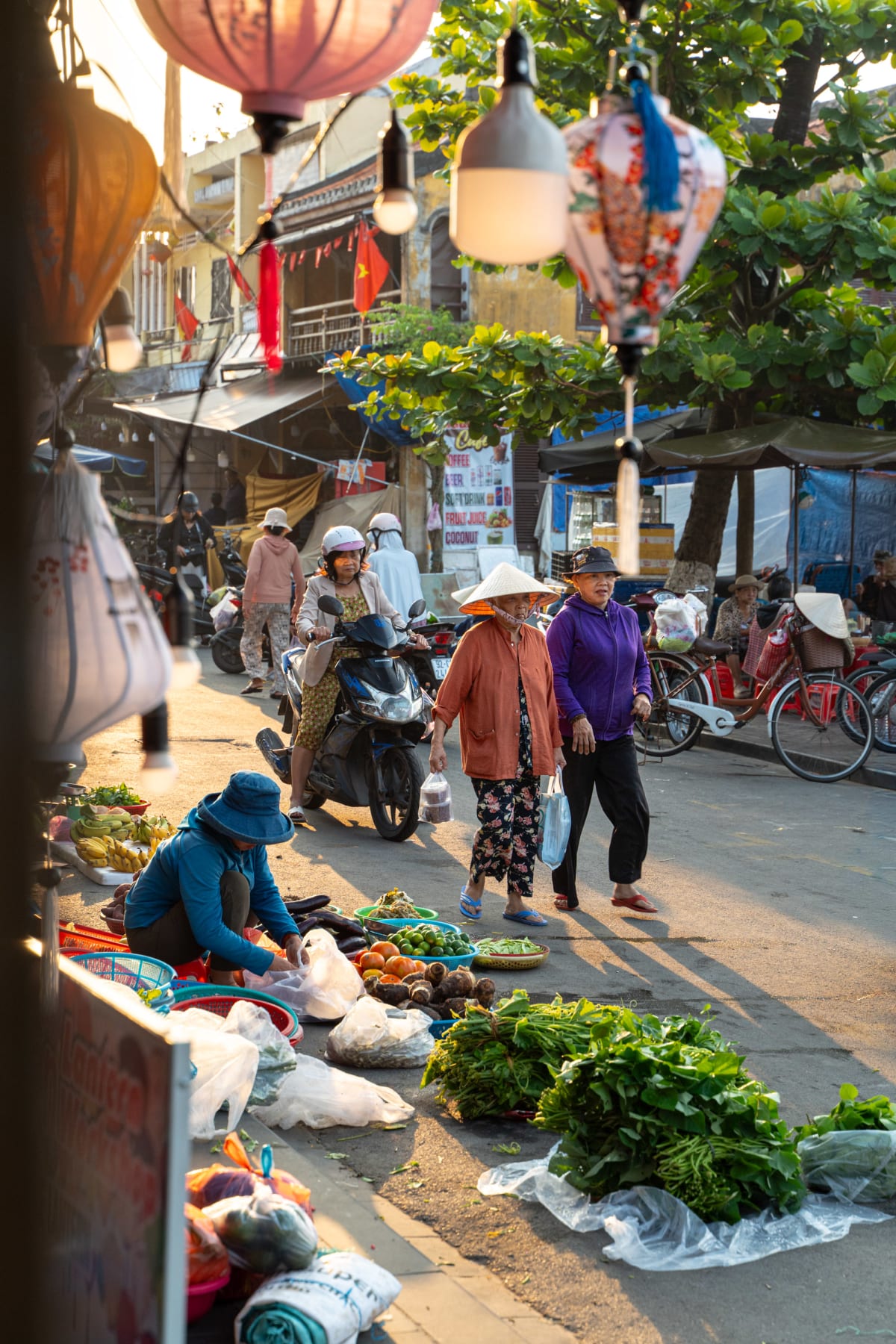 In early morning light, two women walk past a street vendor in a blue bucket hat arranging produce on the pavement, with silk lanterns hanging overhead and a motorbike rider behind them — Pete Walls