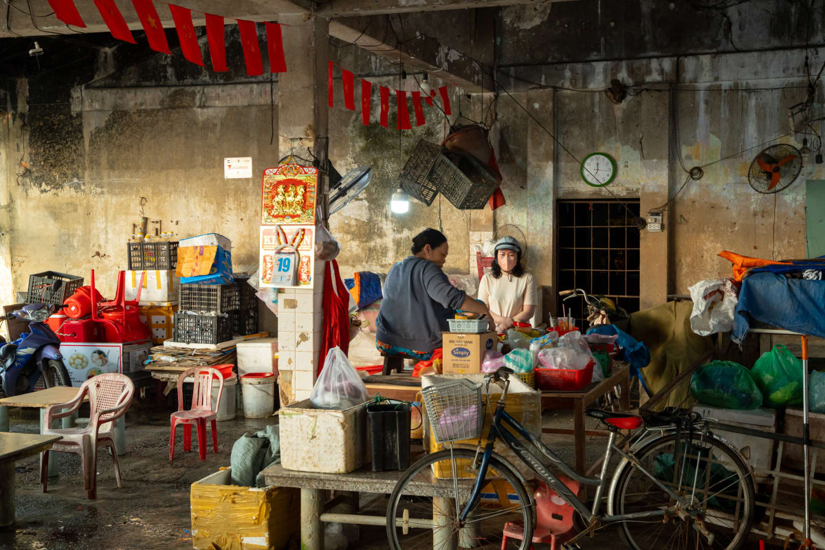 Two women talk across a makeshift counter inside a weathered market hall strung with red flags, surrounded by stacked crates, plastic stools, red chairs, and parked bicycles — Pete Walls