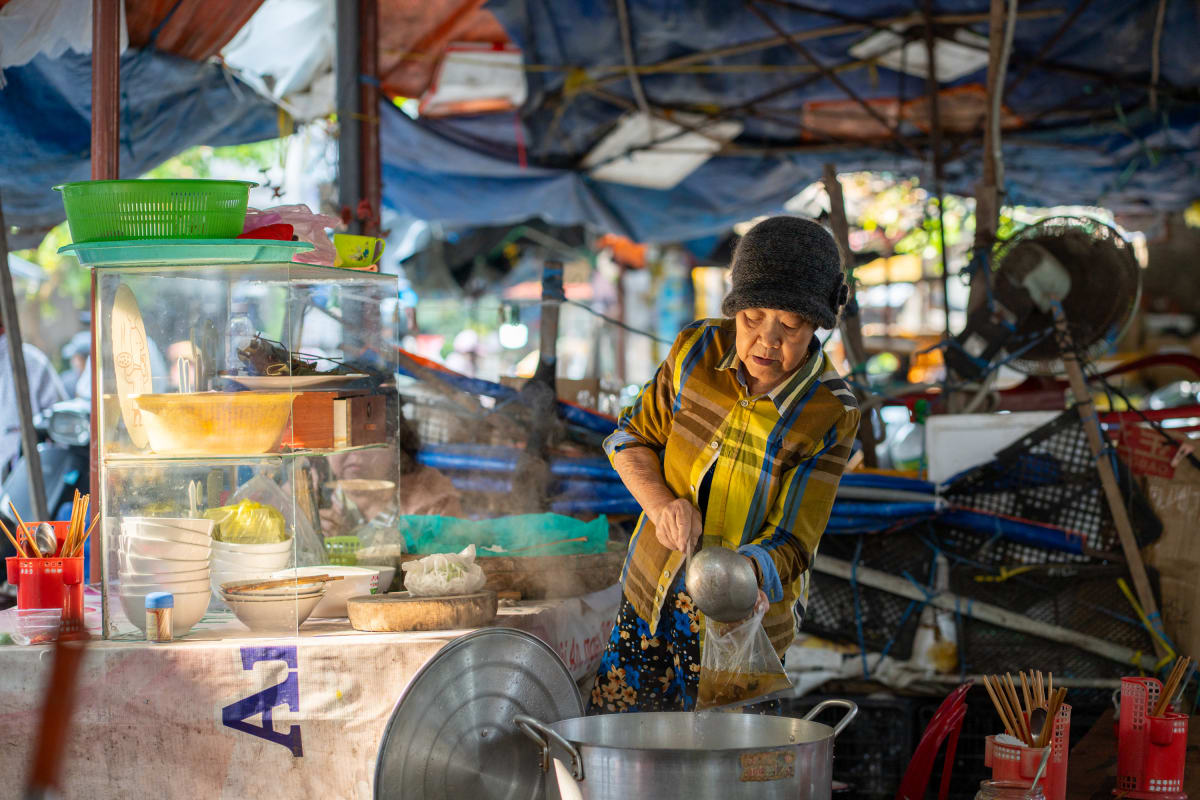 A woman in a knit hat and plaid shirt ladles broth from a large steel pot at a street food stall, beside a glass cabinet stacked with bowls under tarpaulin shade — Pete Walls