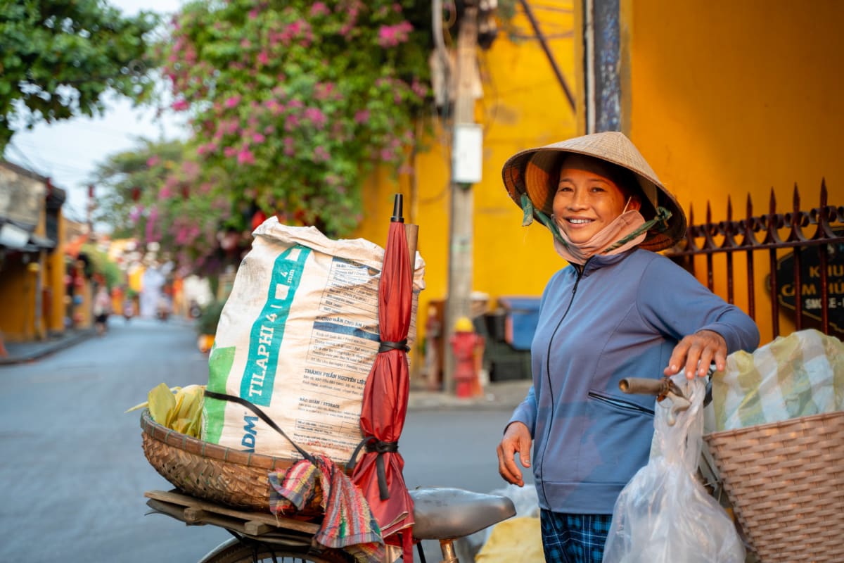 A smiling woman in a conical hat and blue jacket stands with her loaded bicycle — feed sack, furled red umbrella, and baskets — on a street of yellow walls and pink bougainvillea — Pete Walls