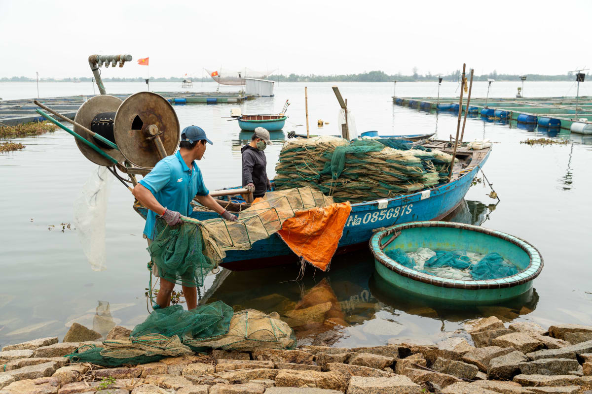 Two men load bundled fishing nets onto a blue wooden boat at a rocky shoreline, one in a turquoise shirt hauling green netting beside a floating round coracle basket — Pete Walls