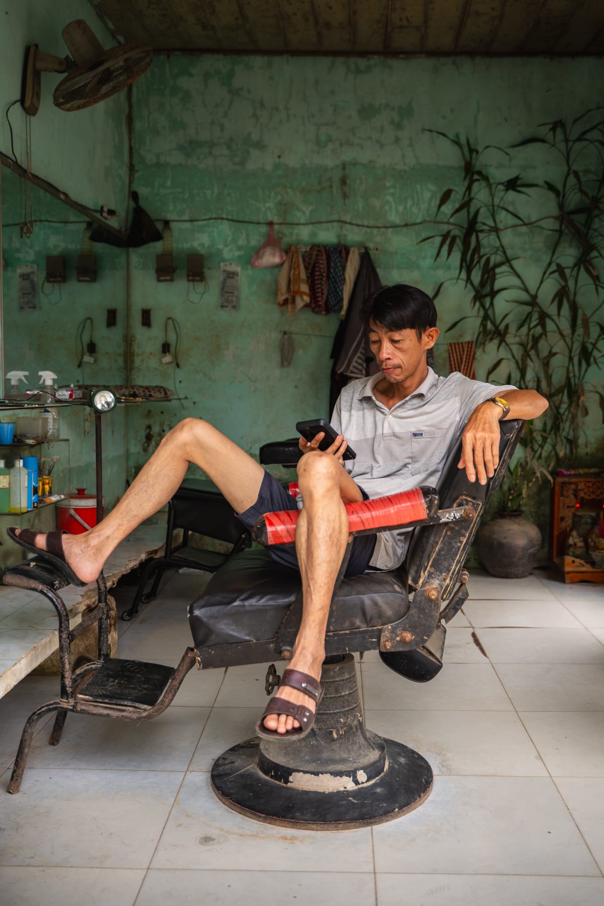 A man in a grey polo reclines in an old barber chair with one leg propped up, looking at his phone inside a green-walled barbershop with bamboo and tools on the wall — Pete Walls