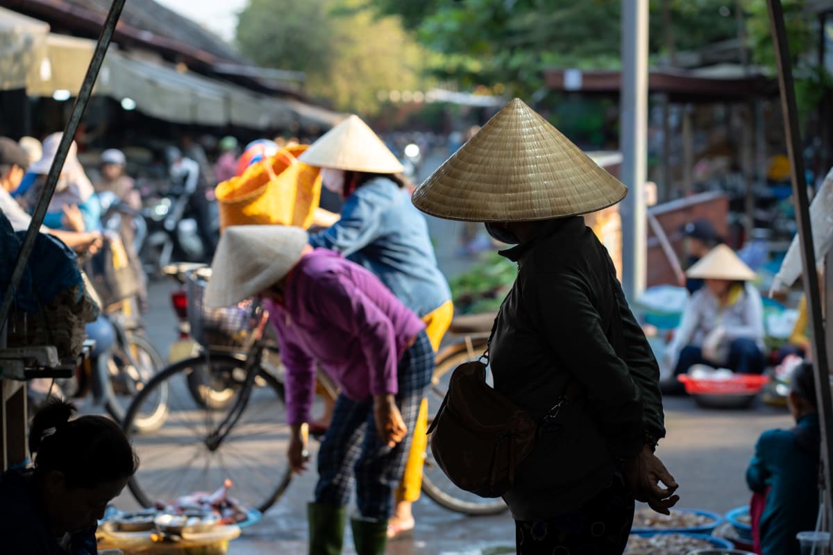 A woman in a conical hat with a shoulder bag stands in market shade as two vendors in purple bend over their stall, with bicycles and shoppers blurred along the lane behind — Pete Walls