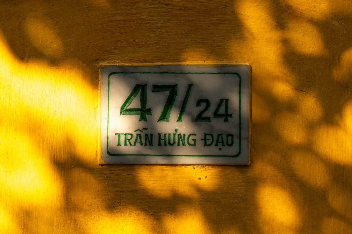 A white house-number plaque reading 47/24 Trần Hưng Đạo mounted on a yellow wall dappled with leaf shadows in warm light — Pete Walls