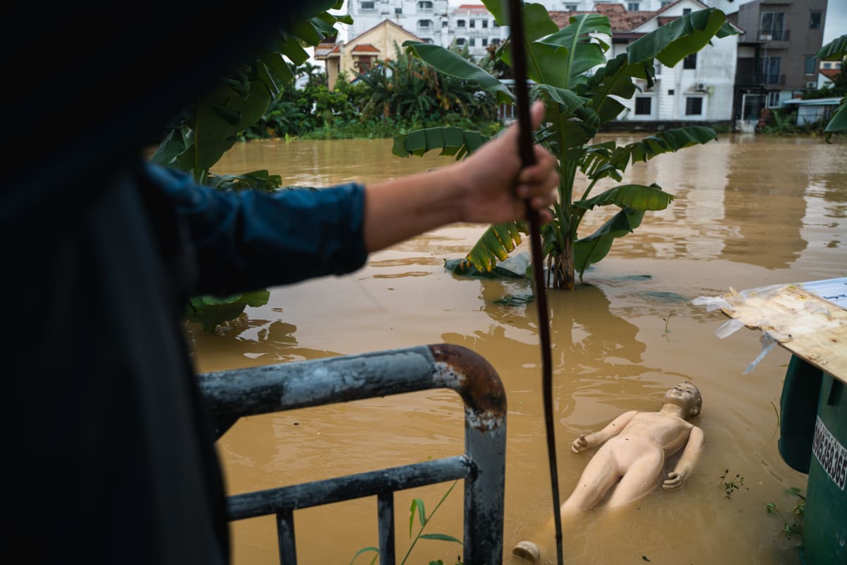 Seen over a man's outstretched arm gripping a pole, a pale mannequin floats on brown floodwater beside a submerged metal gate, banana trees, and houses standing in the flood — Pete Walls