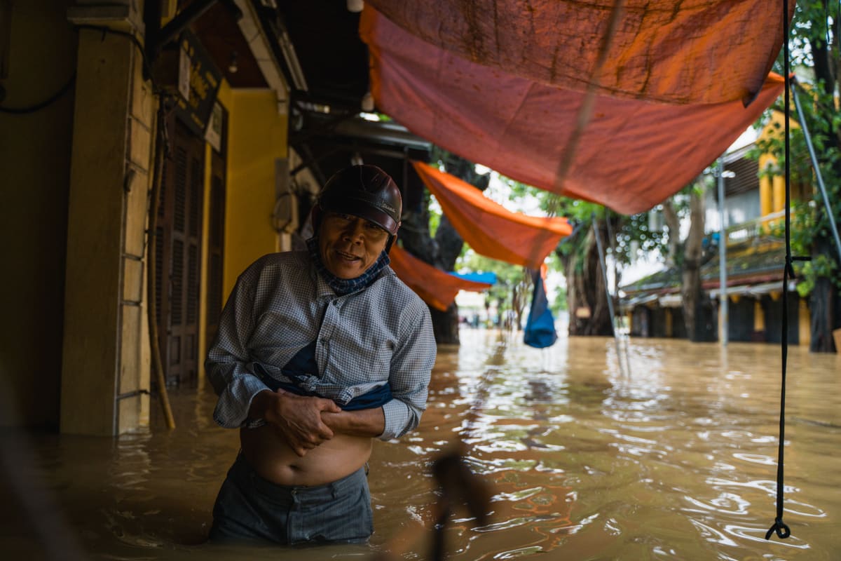 A man in a dark helmet and grey shirt wades waist-deep in muddy floodwater along a shophouse street, clutching his rolled-up shirt beneath sagging orange tarpaulins — Pete Walls