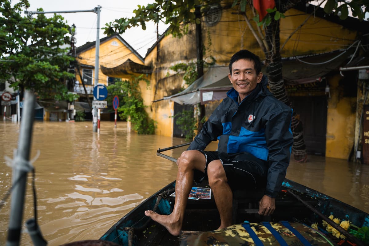A smiling barefoot man in a blue and black jacket sits on the bow of a wooden boat being poled through a flooded street of yellow colonial shophouses — Pete Walls