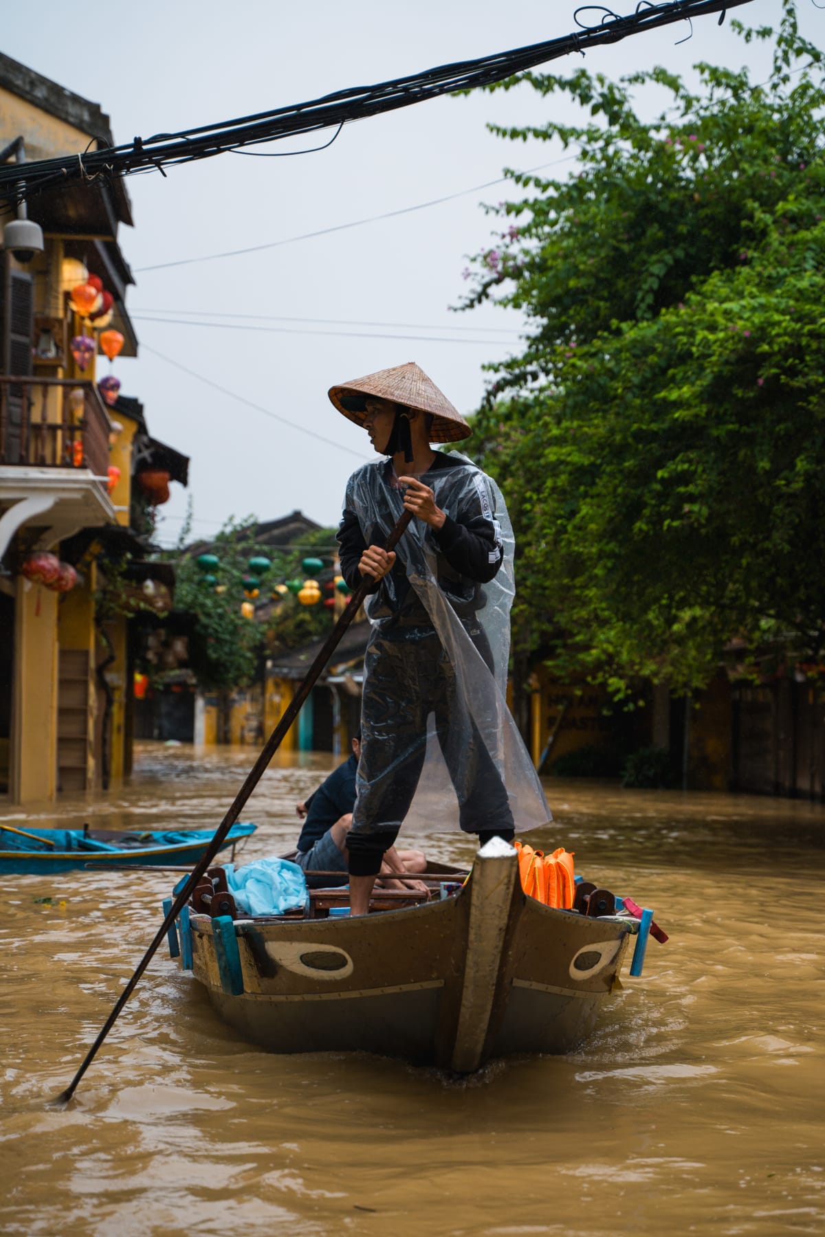 A person in a conical hat and clear plastic rain poncho stands upright in a wooden boat, rowing with a long oar down a flooded lantern-hung street in Hội An's old town — Pete Walls