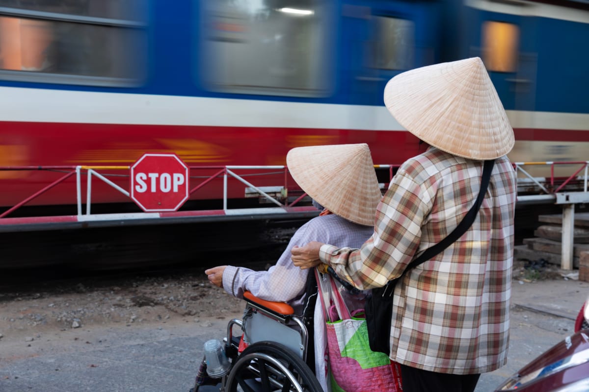 Two women in conical hats wait at a railway crossing behind a red stop sign as a blue-and-red train blurs past, one standing with her hand on her companion’s wheelchair — Paul Evan Green