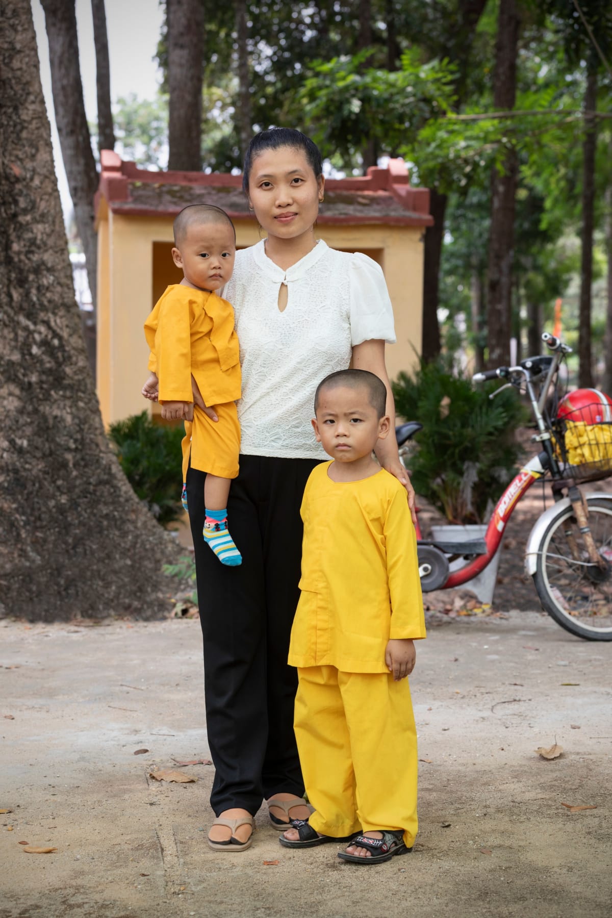 A mother stands with her two young sons dressed in bright yellow, a bicycle parked behind them — Paul Evan Green