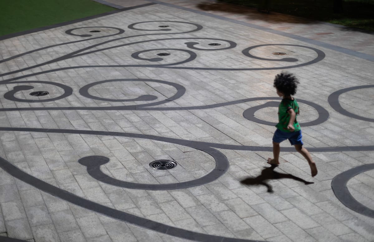 A barefoot child in a green shirt runs at night across a plaza inlaid with dark swirling scrollwork, casting a long shadow — Paul Evan Green