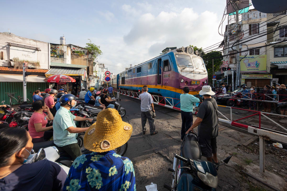 Motorbikes and pedestrians wait at a level crossing as a blue Vietnam Railways locomotive passes through the street — Paul Evan Green