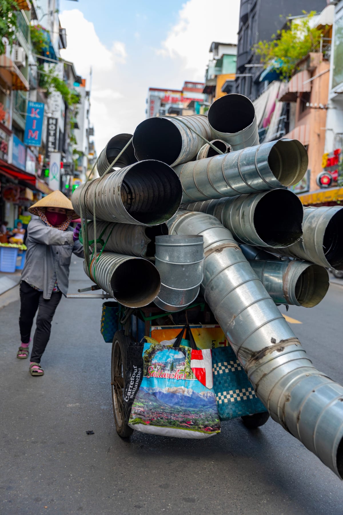 A woman in a conical hat hauls a cart stacked high with galvanized ducting pipes down Bui Vien Street — Paul Evan Green