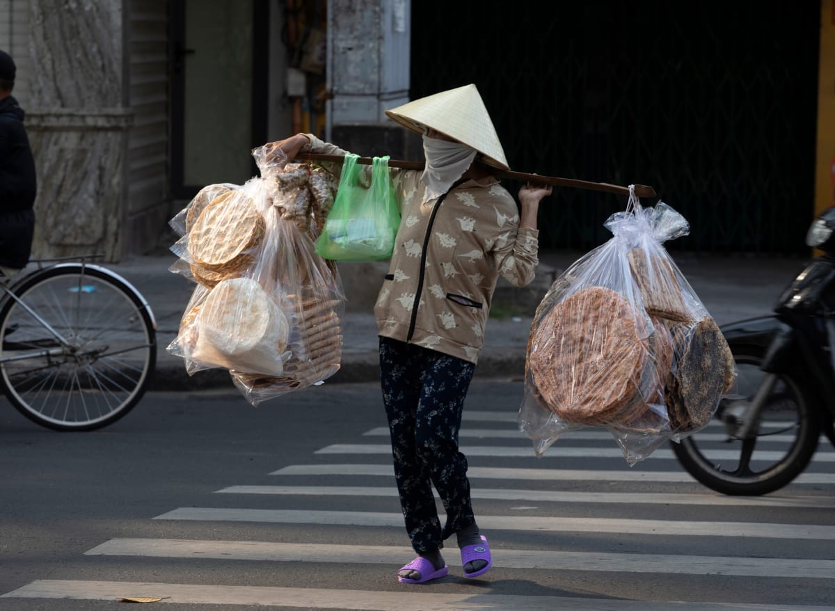 A street vendor in a green conical hat crosses a zebra crossing under a shoulder pole strung with bags of rice crackers — Paul Evan Green
