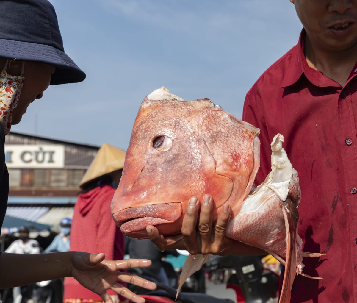 A vendor at a District 8 market holds up a large red snapper head as a customer leans in to inspect it — Paul Evan Green