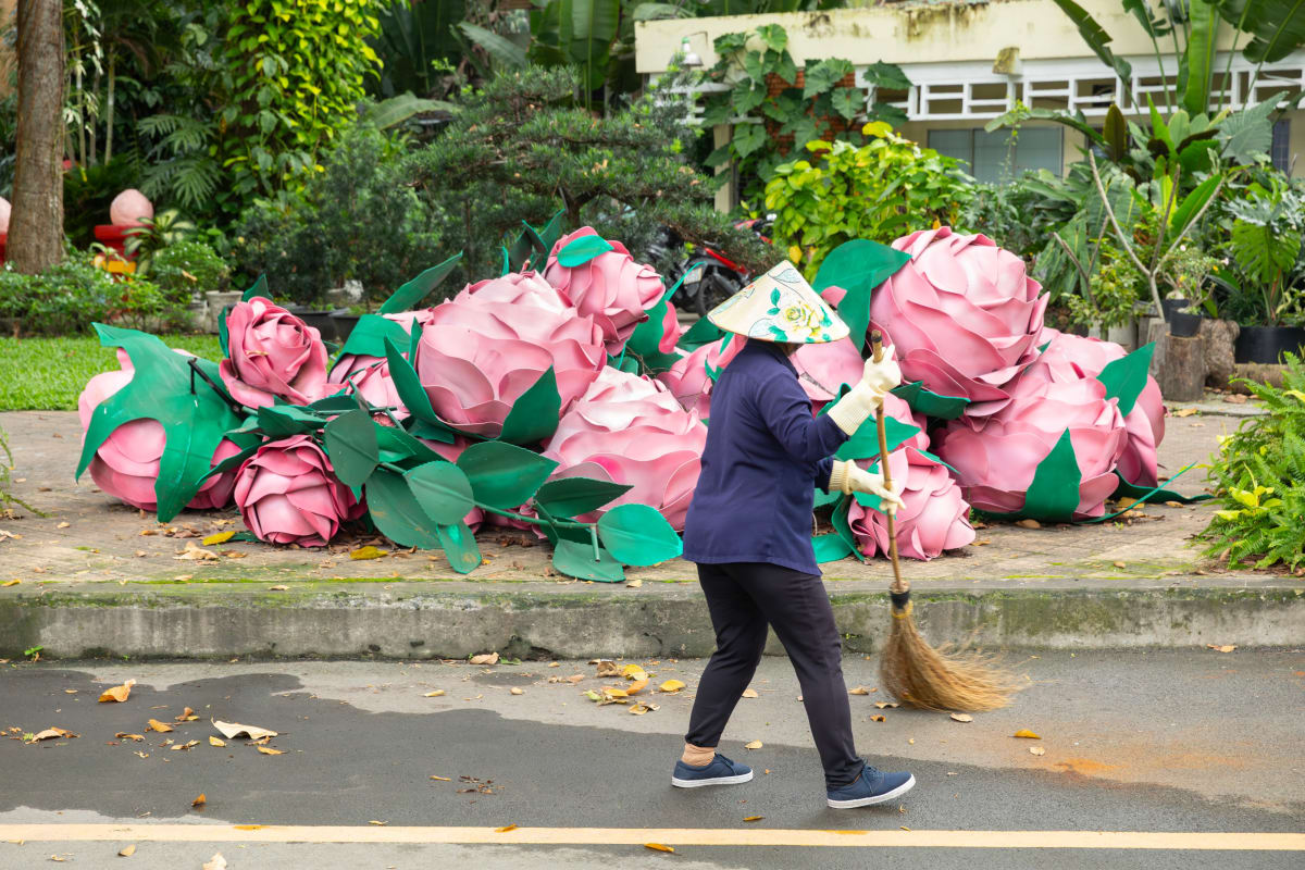 A street cleaner in a painted conical hat sweeps the road past a pile of giant pink rose sculptures — Paul Evan Green