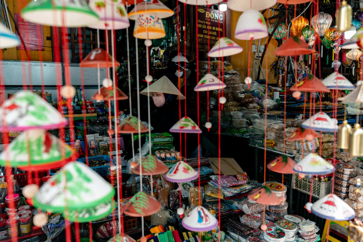 A masked woman in a conical hat sits amid a crowded souvenir stall behind a curtain of dangling miniature painted hats and lanterns — Antonio di Vico