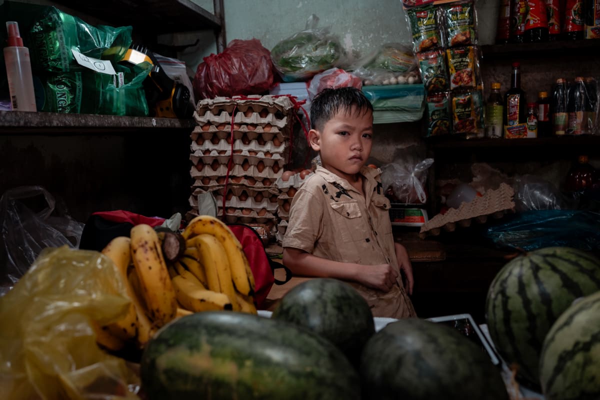 A solemn boy in a beige shirt stands amid stacked egg trays, ripe bananas, and striped watermelons in a dim grocery stall — Antonio di Vico
