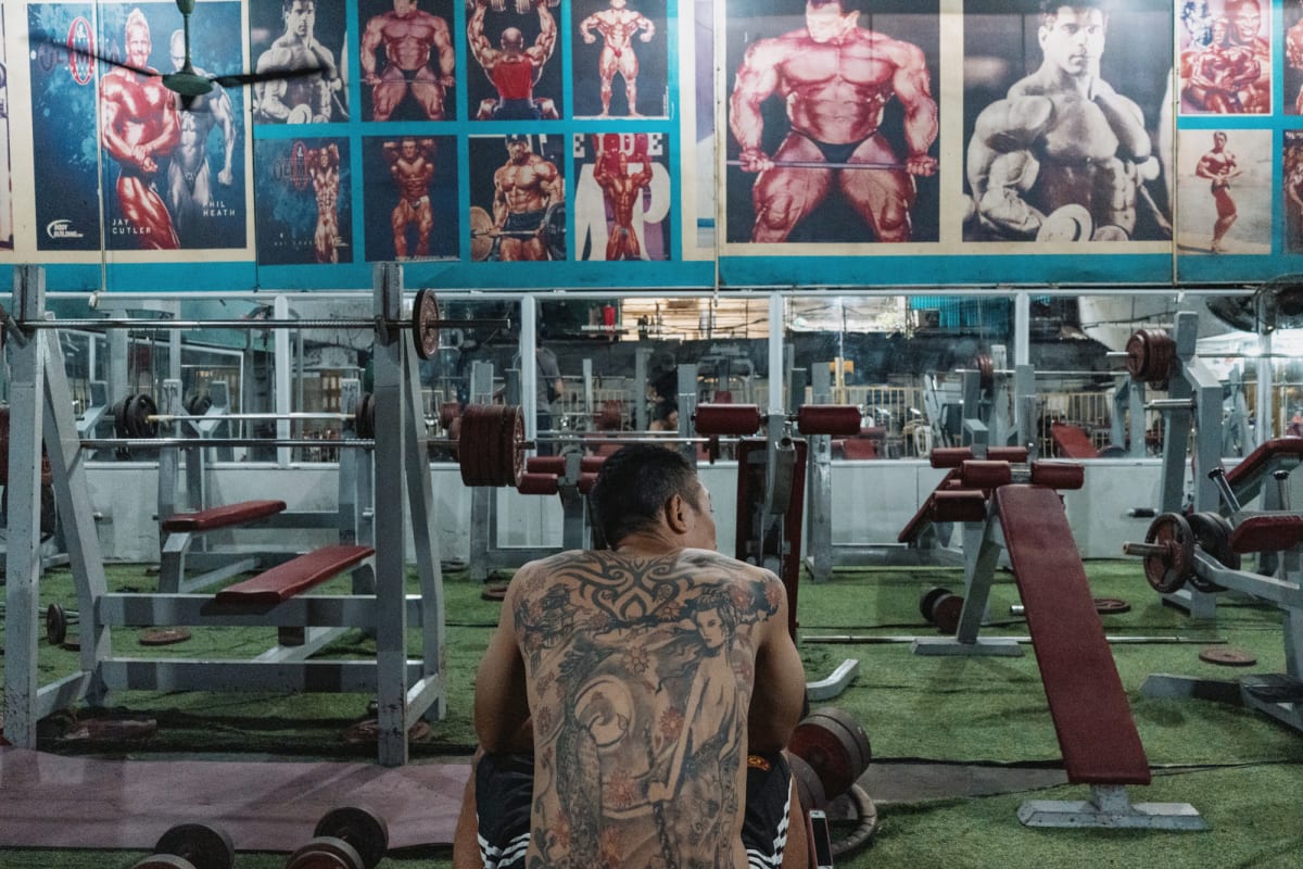 A shirtless man presses two loaded dumbbells overhead on a bench below faded Mr. Olympia bodybuilder posters in a tin-roofed gym — Antonio di Vico