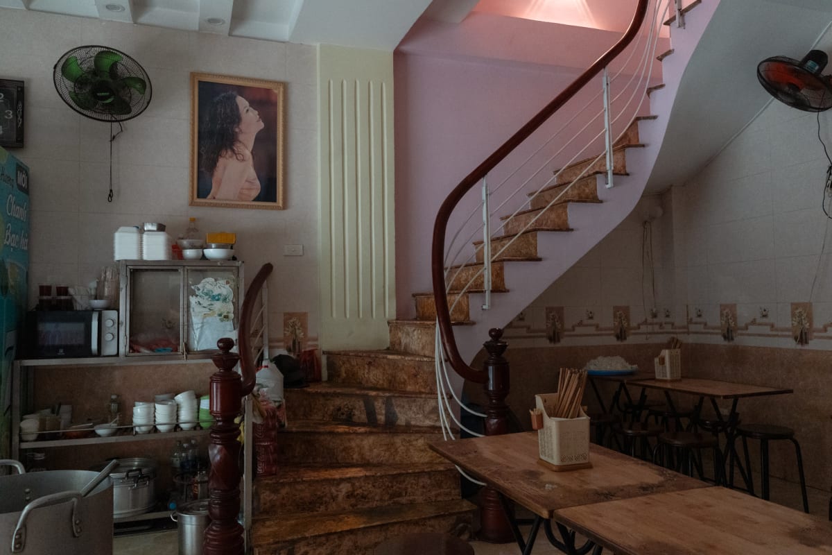 An empty tiled pho shop with a marble spiral staircase, wall fans, stacked bowls, and a framed portrait of a woman — Antonio di Vico