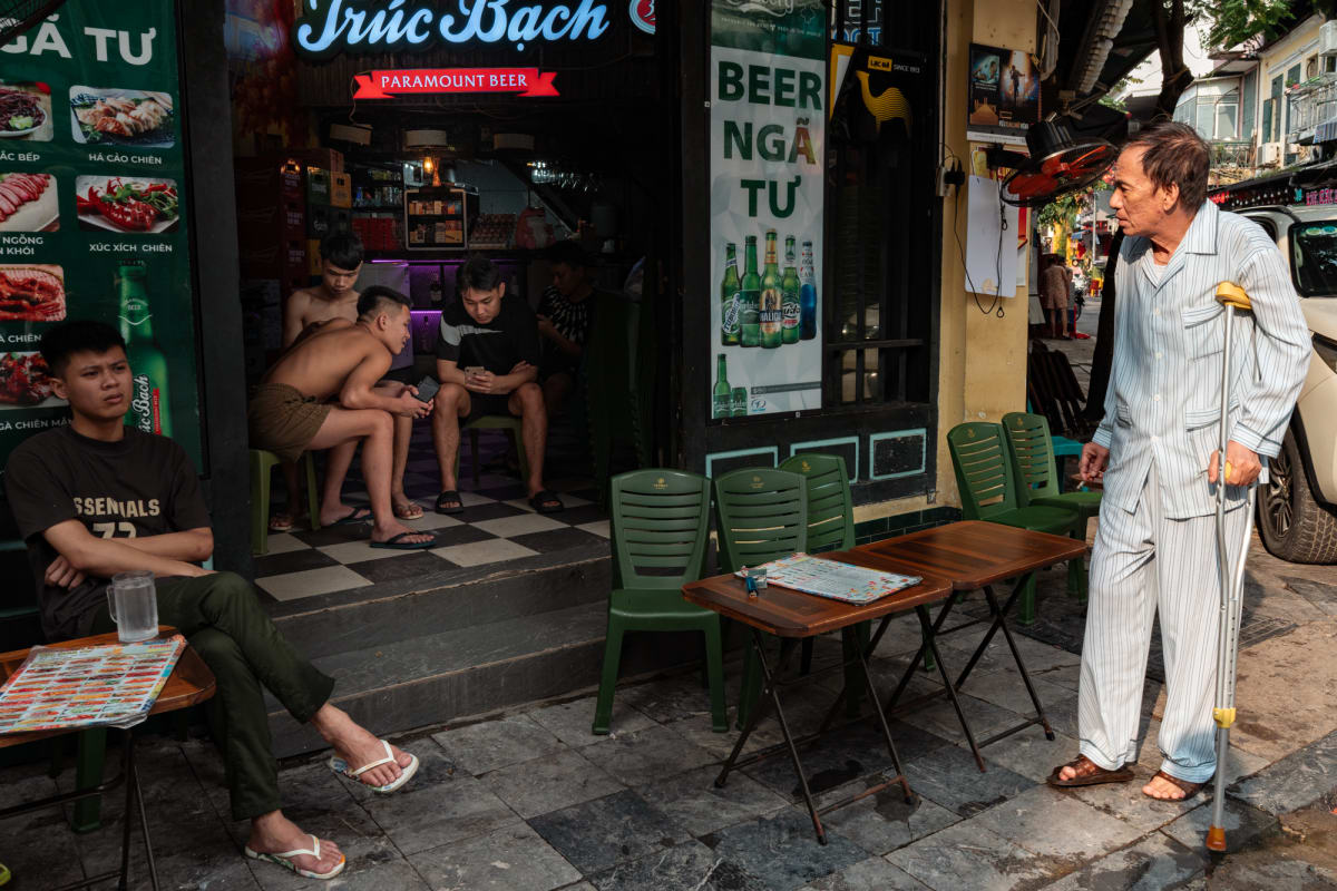 An older man in striped pajamas on a crutch smokes beside a Truc Bach beer bar where shirtless young men sit on green stools — Antonio di Vico