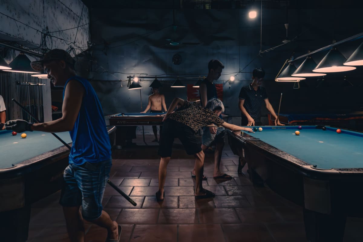 Young men in a dim pool hall lean over glowing blue-felt billiard tables under hanging lamps, one in bright blue walking off — Antonio di Vico